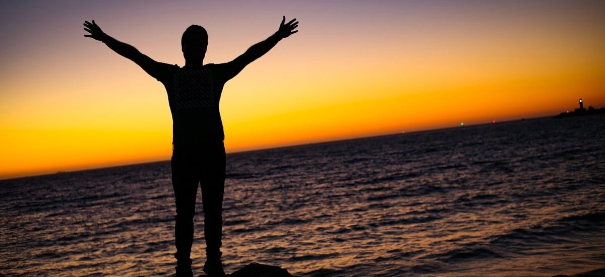A silhouette of a man standing by the sea at sunset, arms raised in a moment of prayer, freedom, and quiet reflection. Warm colors on the horizon create a peaceful and spiritual atmosphere.