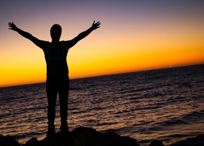 A silhouette of a man standing by the sea at sunset, arms raised in a moment of prayer, freedom, and quiet reflection. Warm colors on the horizon create a peaceful and spiritual atmosphere.